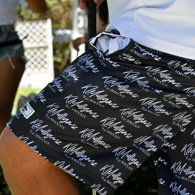 A close-up shot of the Legacy Shorts from KLMulliganz, prominently displaying the brand’s signature logo in an all-over print. The image captures the intricate detail and high-quality fabric of these black shorts, which are designed for both golf enthusiasts and those embracing a lifestyle statement. The Legacy Shorts combine comfort and style, making them perfect for the course or casual wear. The background subtly features another golfer, emphasizing the versatility and broad appeal of the KLMulliganz bra